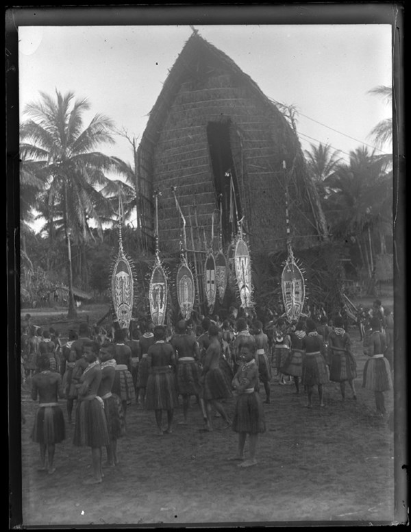 A group of Elema people, standing in front of a thatched roof men's house, all wearing plant fibre skirts, neck ornaments, and nose ornaments, with several people holding hevehe (masks); Purari Delta, Papua New Guinea. © The Trustees of the British Museum (CC BY-NC-SA 4.0)