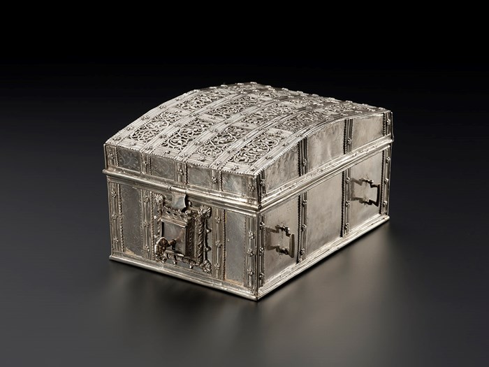 Metal cuboid casket with ornate design on the lid, handles on the outside and a large key in a large lock on one end