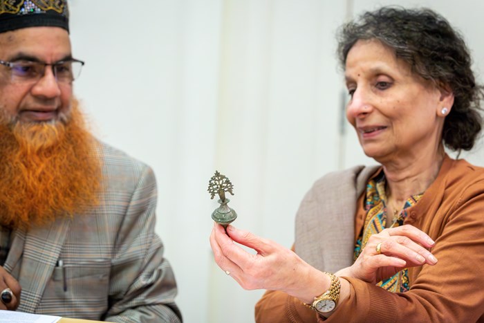 Two older people look at a small, elaborately decorated pot made of metal together. The top of the pot is shaped like a tree. The man wears an embroidered black hat and has a beard. The woman holds the pot in her hand.