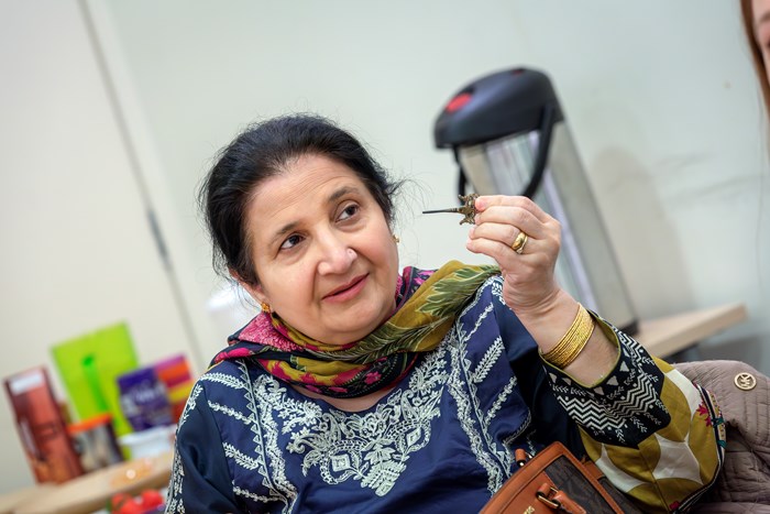 An older woman wearing a colourful scarf and embroidered top with dark hair holds the lid and applicator from a kohl pot up to her eyes, to demonstrate how kohl was applied.