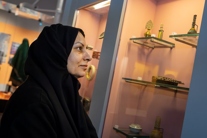 A profile of a woman wearing a black hijab (material covering over her head and shoulders) intently looks at golden and other small objects from west and central Asia.