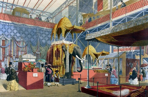 A colour drawing of the Indian pavilion at the Great Exhibition of 1851,
showing people in Victorian dress standing in a large glass building surrounded by
colourful swathes of textiles and objects, including an elephant adorned in elaborate
materials with a golden, domed structure on top.