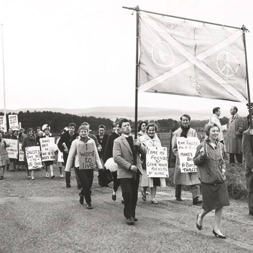 Black and white photo of a large group of young people walking and holding homemade signs with peaceful slogans.