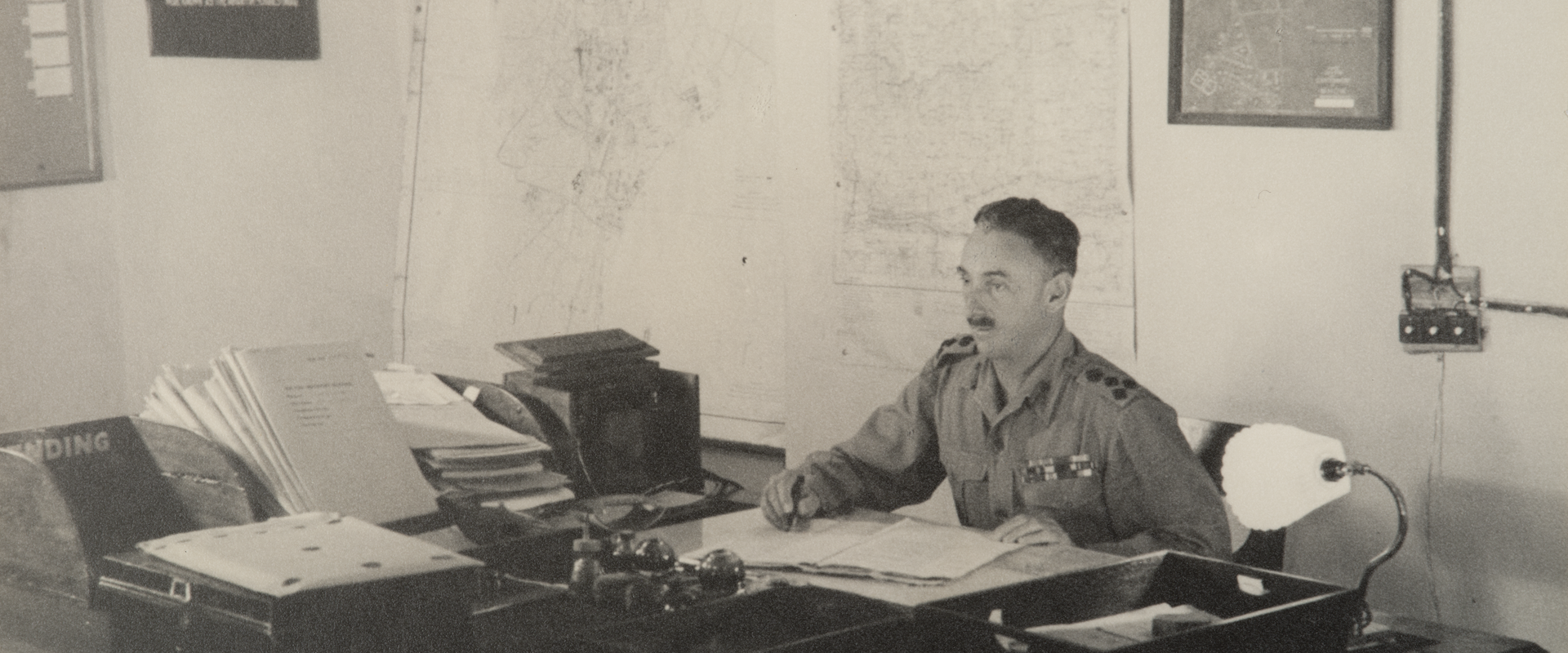 Brigadier Norman Macdonald sits at a desk surrounded by papers.