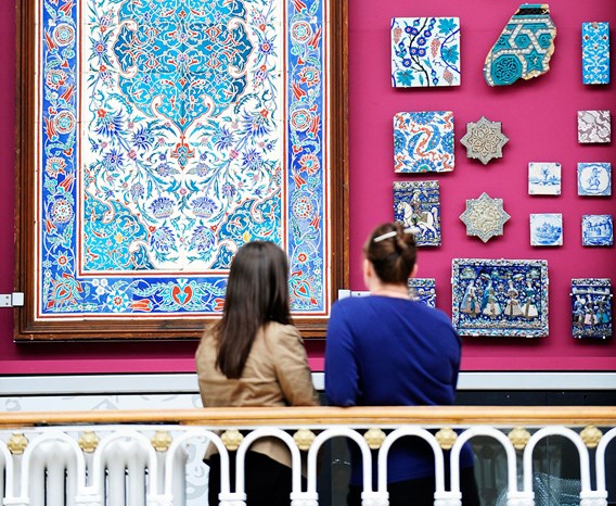 Two visitors looking at mosaic tiles hanging on a wall.