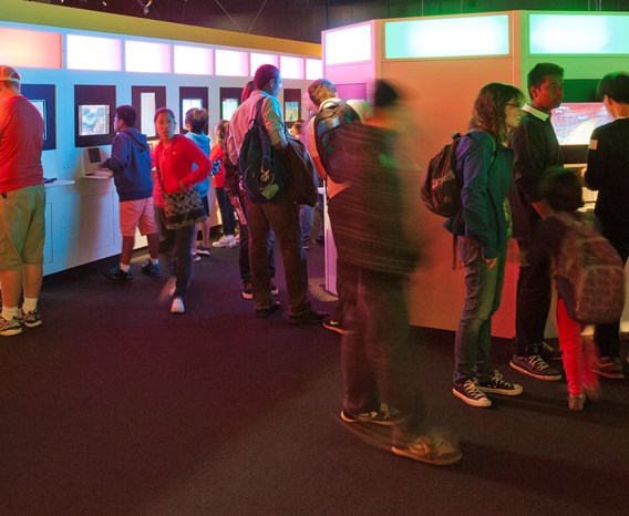 Ten players crowd around a line of consoles in an exhibition space at Game On in Ontario Science Centre.ario Science Ce