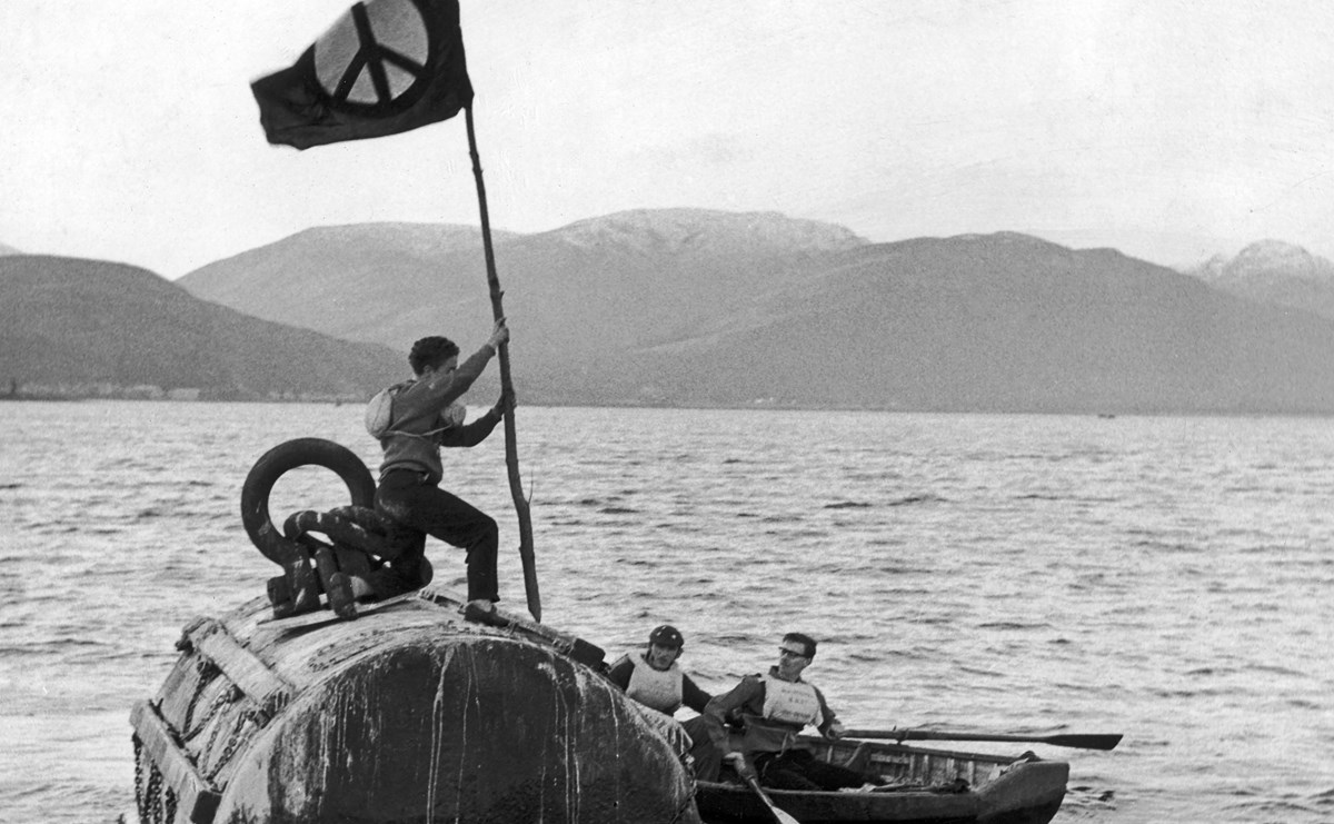 A protestor holding a peace flag stands on top of a submarine as a police boat comes in alongside the vessel. 