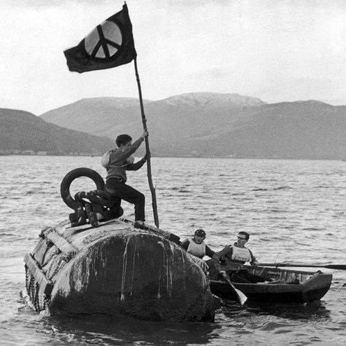 A black and white photo of a person holding a flag who is standing on a submarine which is at the surface of a loch.