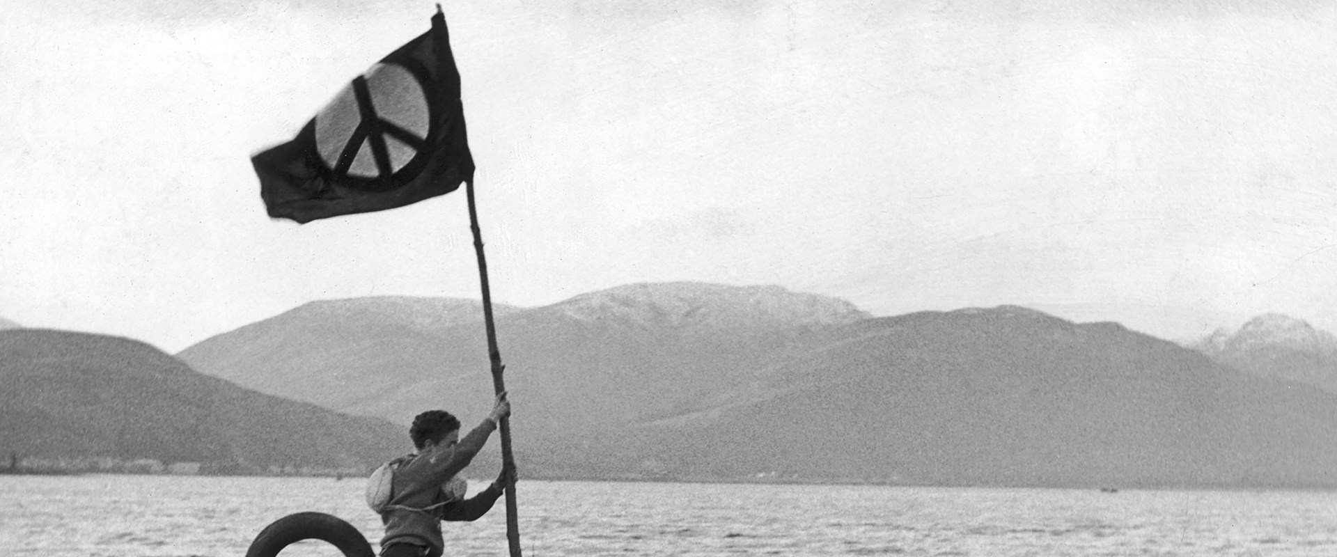 A black and white photo of a person holding a flag who is standing on a submarine which is at the surface of a loch.