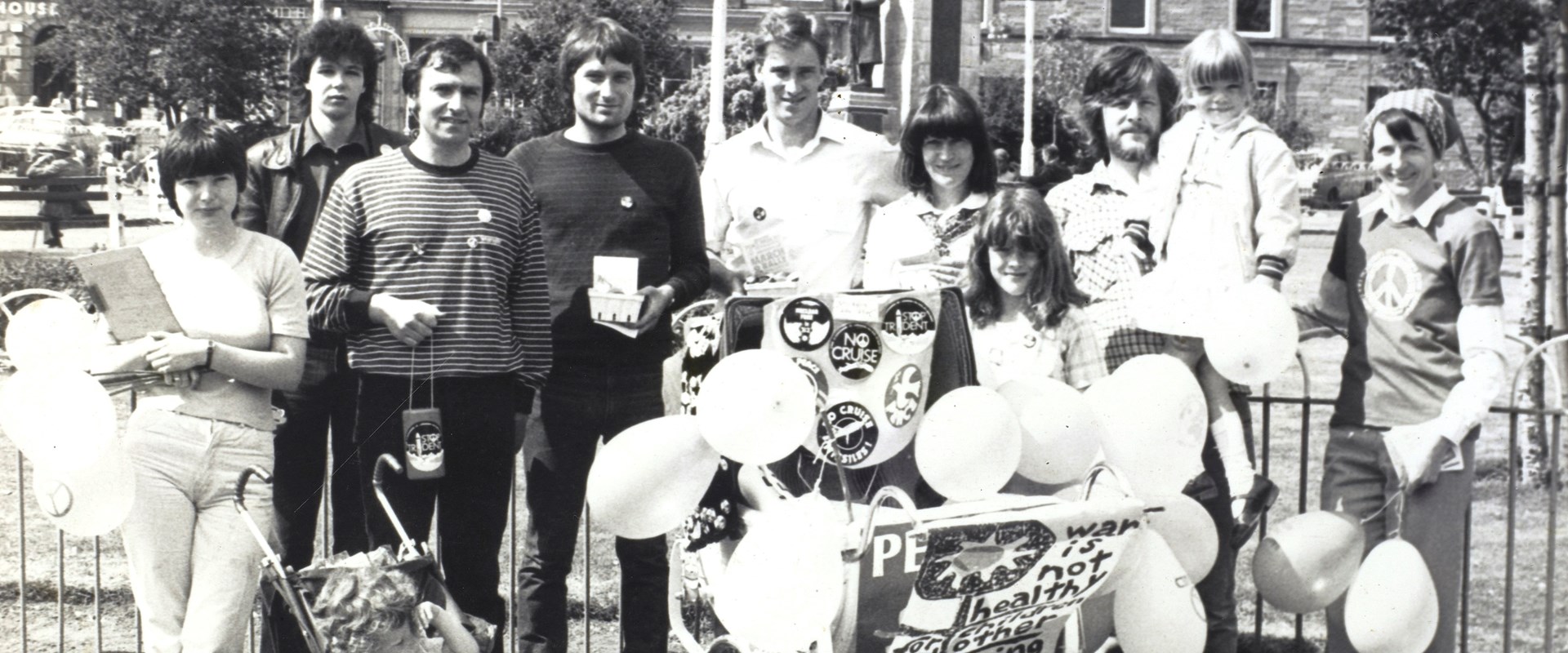 A black and white photo of a group of people standing outside with a cart with balloons.
