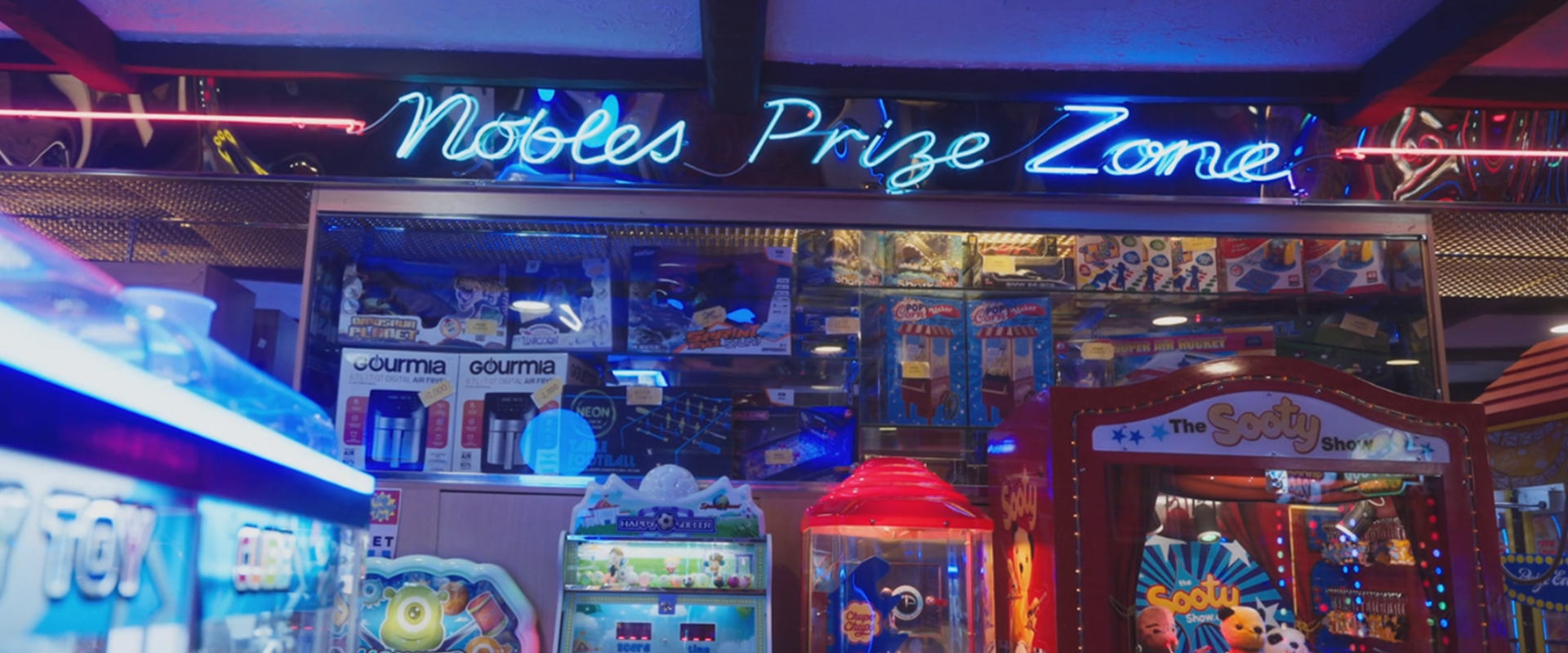 The inside of Nobles Amusements, showing a backdrop of arcade games against a neon blue sign that says 'Nobles Prize Zone'